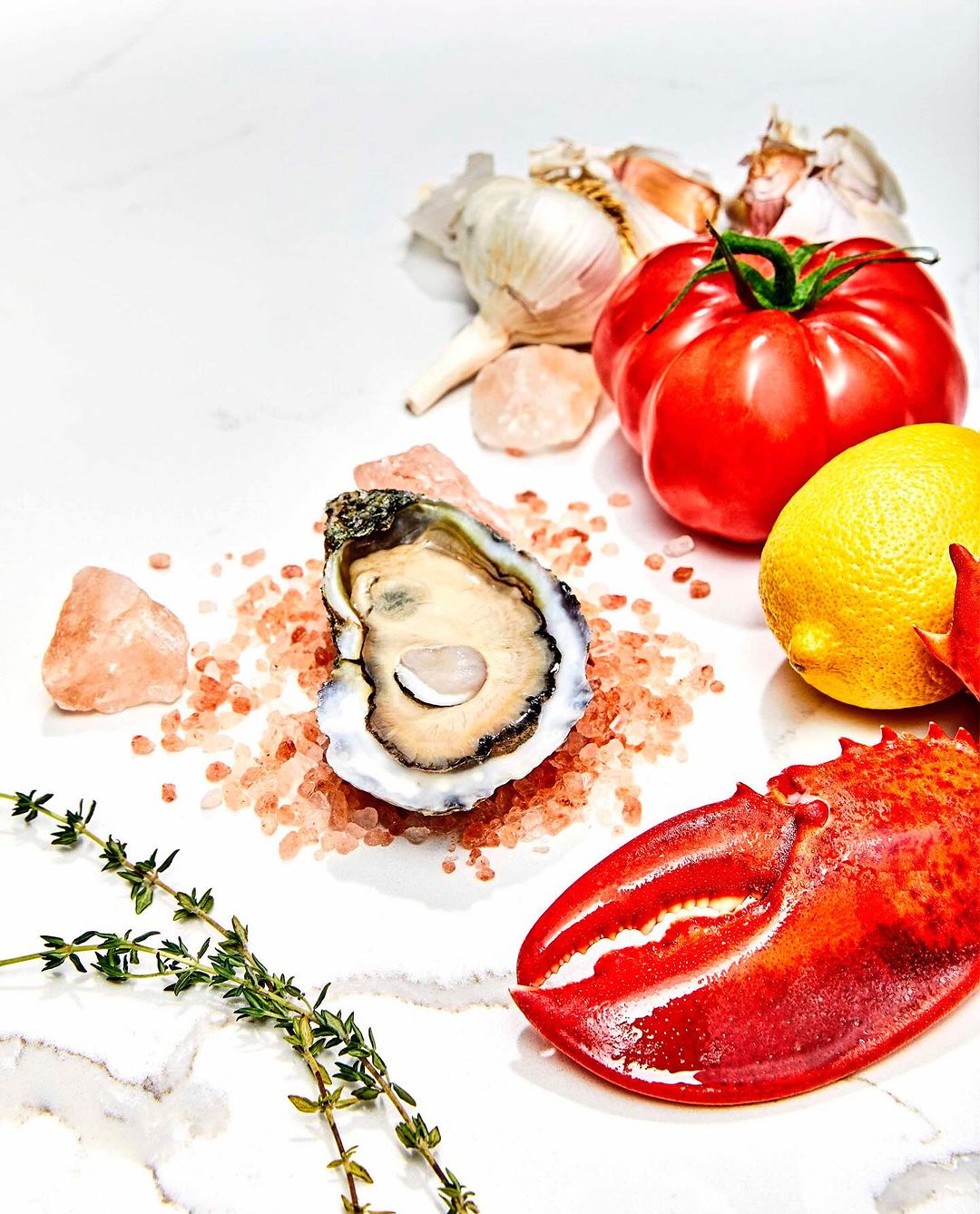 Overhead shot of an oyster, lobster claw, tomato, garlic, lemon, and thyme sprigs on a white marble surface, creating a fresh, vibrant culinary scene.