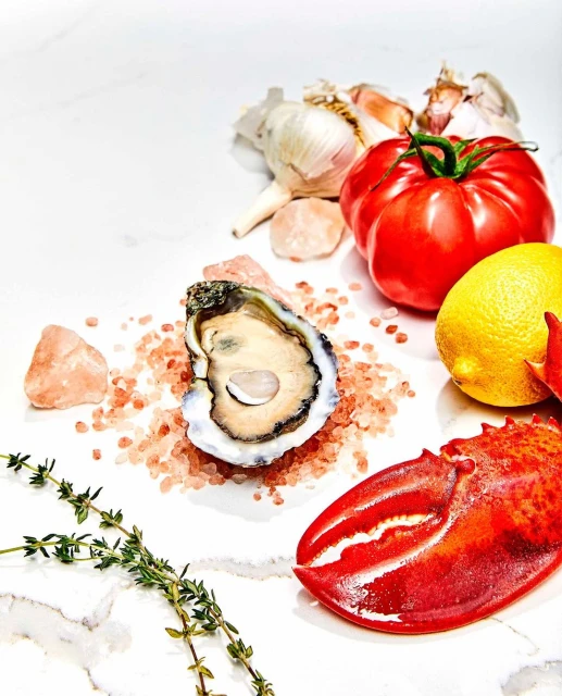 Overhead shot of an oyster, lobster claw, tomato, garlic, lemon, and thyme sprigs on a white marble surface, creating a fresh, vibrant culinary scene.