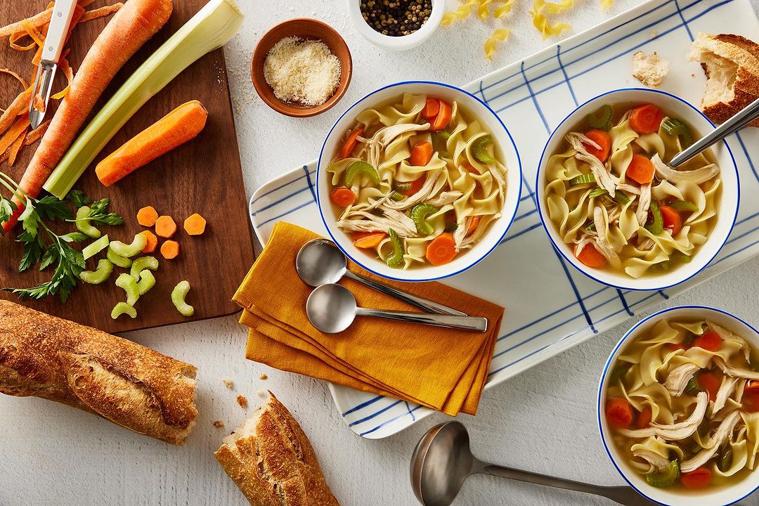 Overhead shot of chicken noodle soup in bowls with carrots, celery, noodles, and chicken, complemented by bread and fresh vegetables.