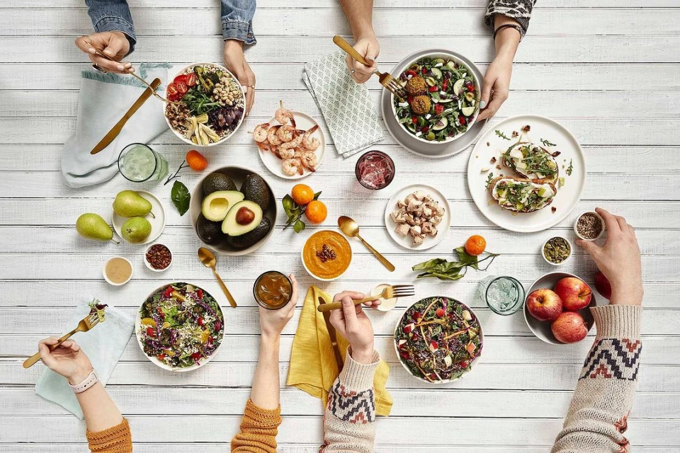 Overhead shot of a white wooden table with food bowls, plates of appetizers, fruit, and beverages being enjoyed by several people.
