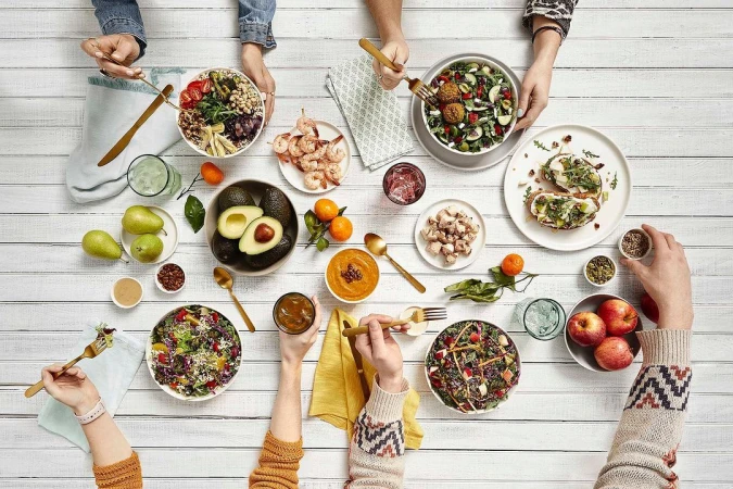 Overhead shot of a white wooden table with food bowls, plates of appetizers, fruit, and beverages being enjoyed by several people.