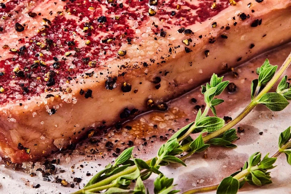 Close-up on a seasoned raw steak with visible marbling, sprinkled with salt and pepper, complemented by fresh thyme sprigs.