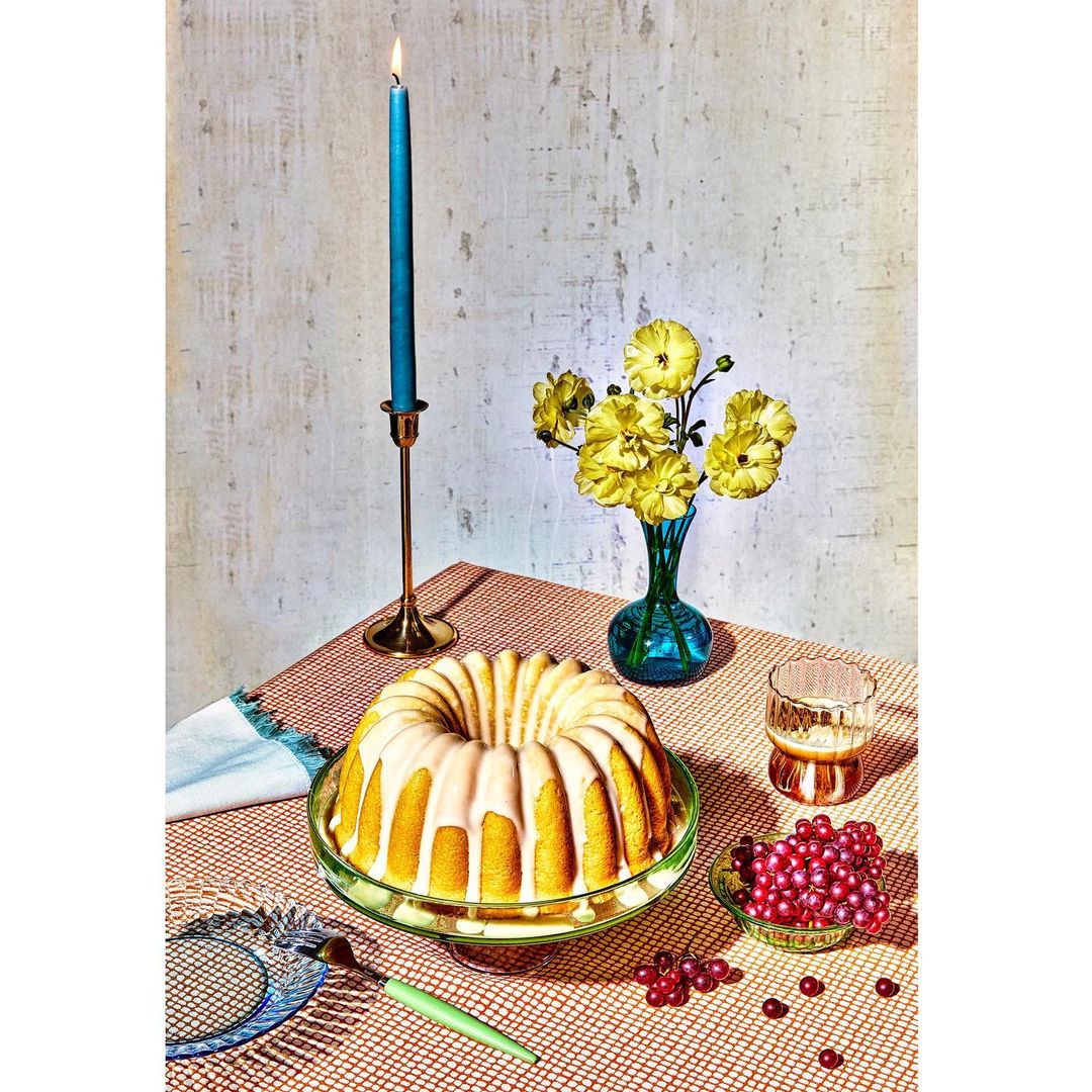 Golden bundt cake with icing on a green cake stand, accompanied by a candle, flowers, glass, berries, and silverware on a patterned tablecloth.