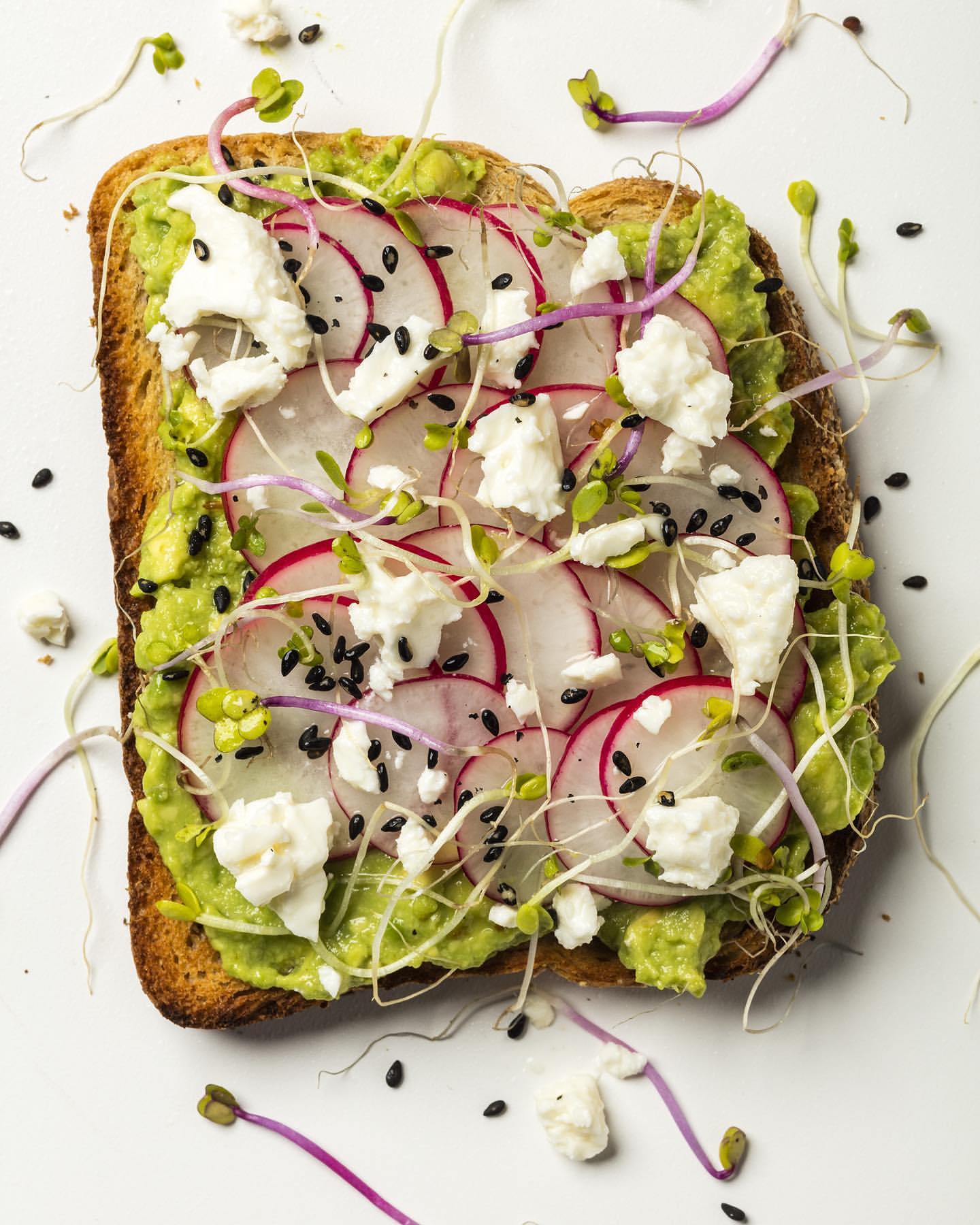 Overhead shot of toast with avocado, radish slices, crumbled cheese, and sprouts on a white surface, prepared food photography.