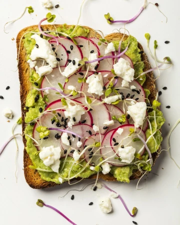 Overhead shot of toast with avocado, radish slices, crumbled cheese, and sprouts on a white surface, prepared food photography.