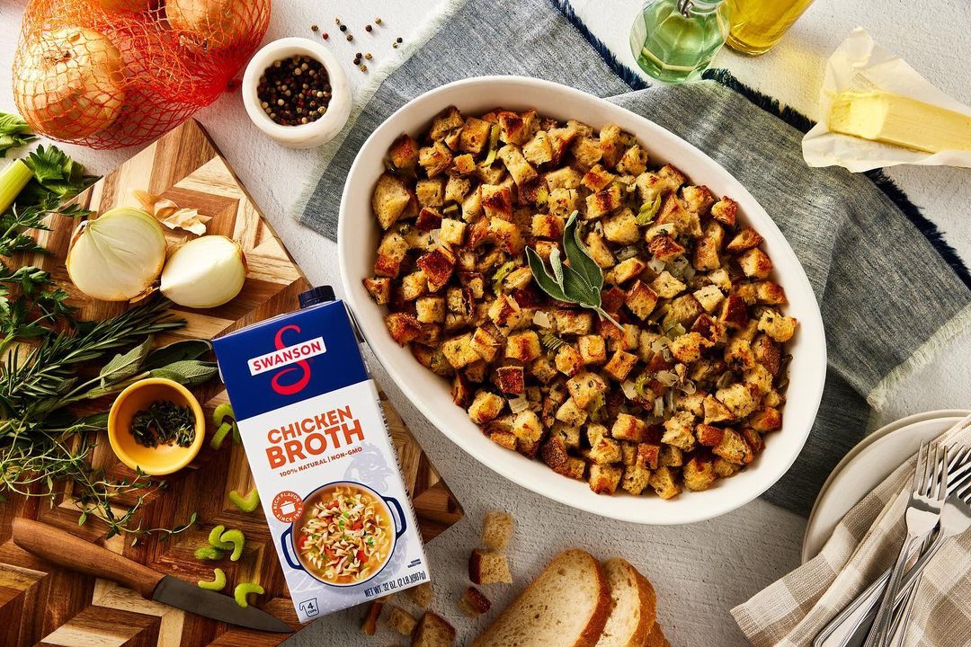 Overhead shot of stuffing in a white bowl, Swanson chicken broth, onions, herbs, silverware, butter and olive oil on a gray surface.