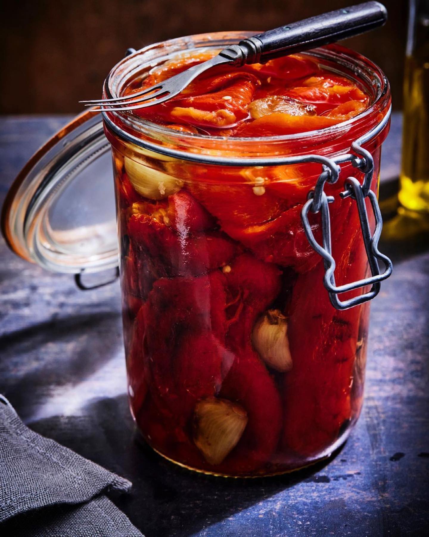 Open glass jar filled with pickled red peppers, topped with garlic cloves and a fork, sitting on a dark tabletop with a grey cloth.