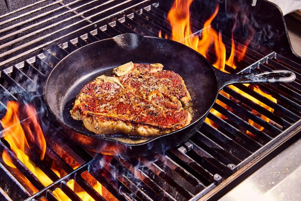 A cast iron pan sits on a flaming grill, cooking a seasoned steak. The flames surround the pan and lick at the meat.