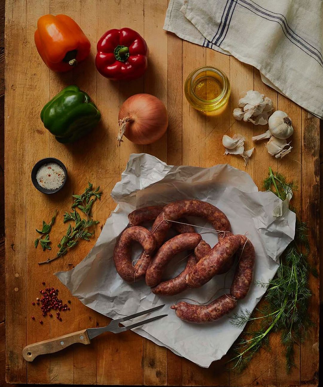 An overhead shot displays uncooked sausages on parchment paper, surrounded by bell peppers, an onion, garlic, herbs, and a meat fork on wood.