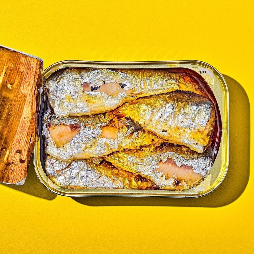 Overhead shot of tinned sardines in olive oil with the lid ajar on a bright yellow background in a studio setting with natural light.