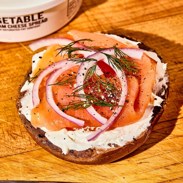 Open-faced bagel sandwich with cream cheese, salmon, red onions, and dill on a wooden board with a container of cream cheese.