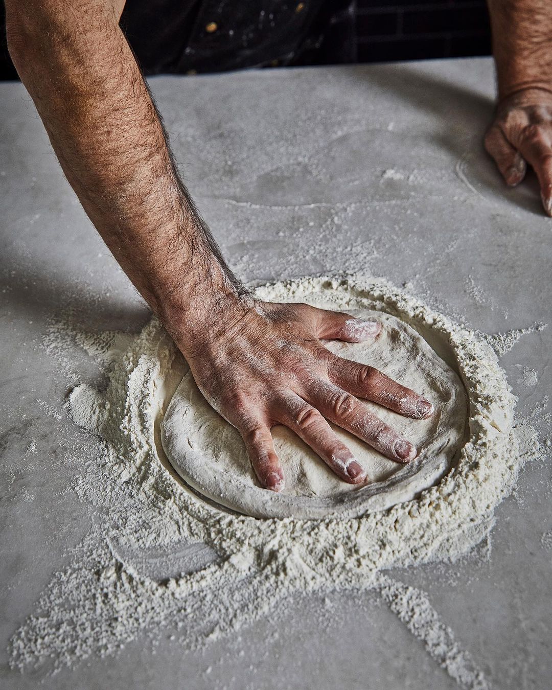 Man's hand presses down on a round mass of dough encircled by flour, set on a grey surface in a softly illuminated indoor setting.