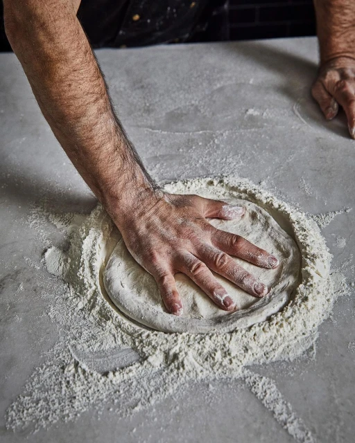 Man's hand presses down on a round mass of dough encircled by flour, set on a grey surface in a softly illuminated indoor setting.