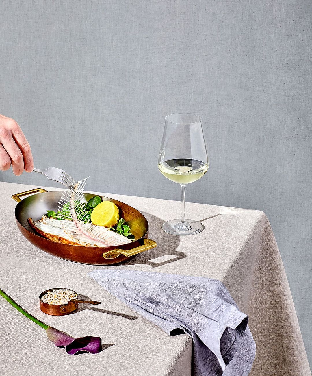 Still life shot of fish skeleton, lemon, and herbs in a metal pan alongside white wine and a cloth napkin on a table.