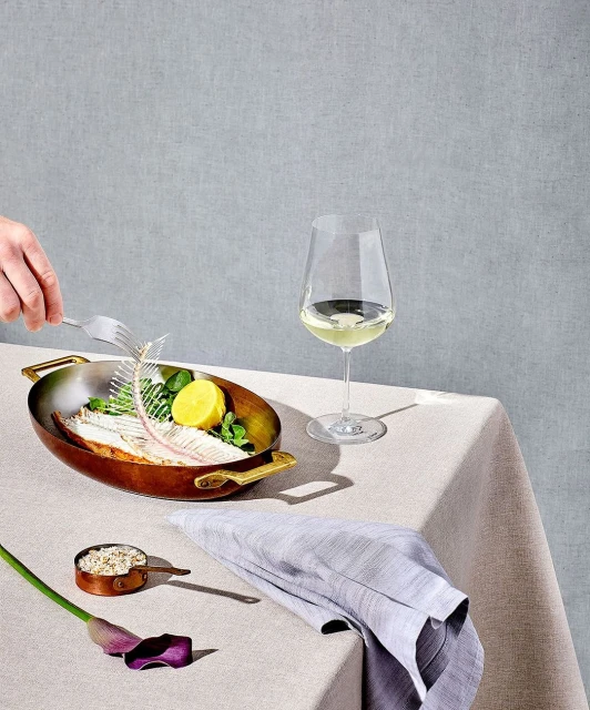 Still life shot of fish skeleton, lemon, and herbs in a metal pan alongside white wine and a cloth napkin on a table.