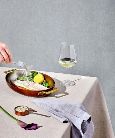 Still life shot of fish skeleton, lemon, and herbs in a metal pan alongside white wine and a cloth napkin on a table.