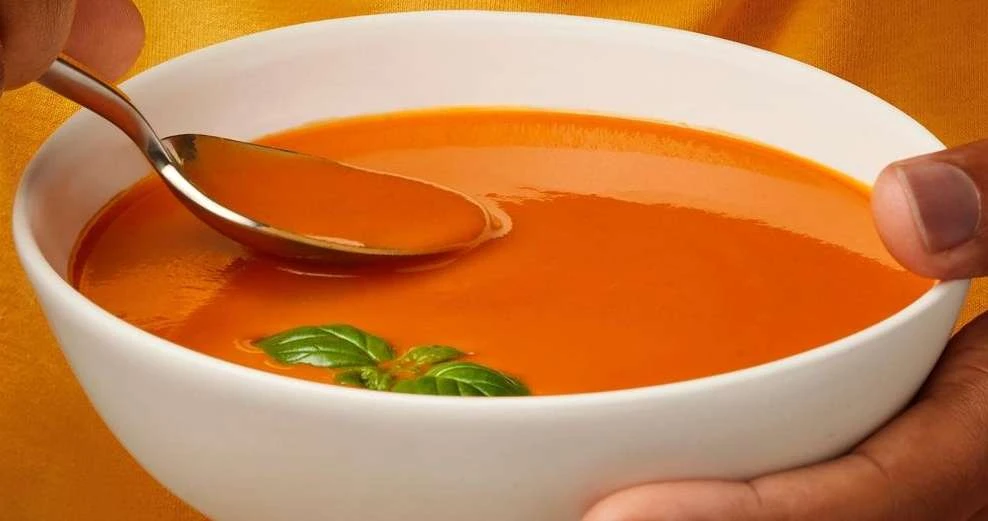 Bowl of tomato soup held by a person with a spoon inside and garnished with basil, presented on a yellow background, close-up shot.