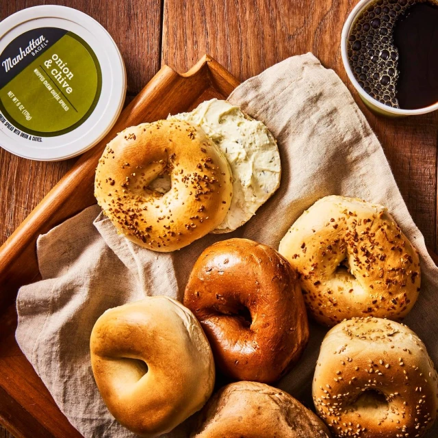 Overhead shot of bagels and cream cheese on a wooden tray with a linen cloth, alongside a container of spread and a cup of coffee.