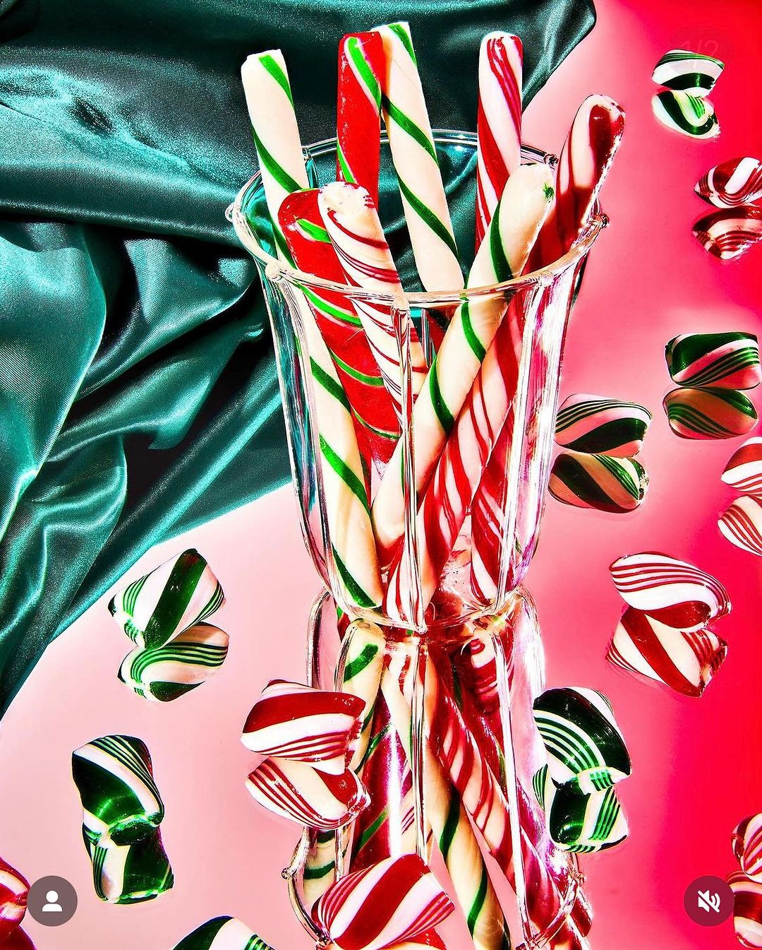 Close-up of candy canes in a glass vase on a pink surface with a green fabric backdrop, creating a festive, colorful holiday theme.