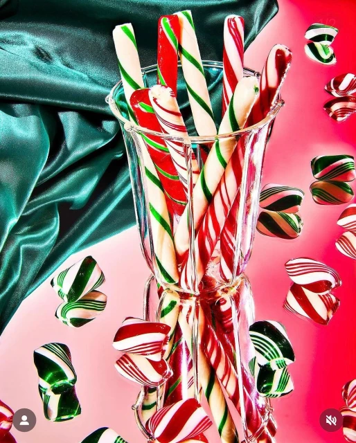 Close-up of candy canes in a glass vase on a pink surface with a green fabric backdrop, creating a festive, colorful holiday theme.