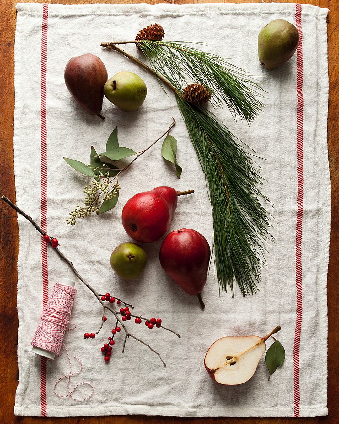 Overhead view of red and green pears, pine sprigs with cones, winterberries, and string on a white linen towel with red stripes.