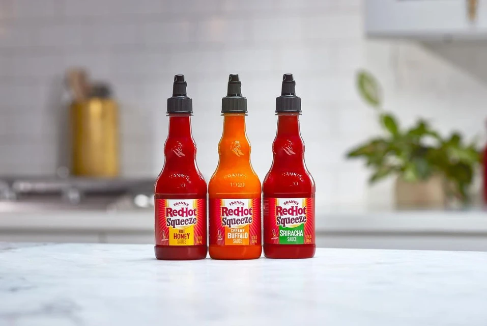 Three Frank's RedHot Squeeze bottles in a row: Hot Honey, Creamy Buffalo, and Sriracha, sitting on a marble counter in a bright kitchen.