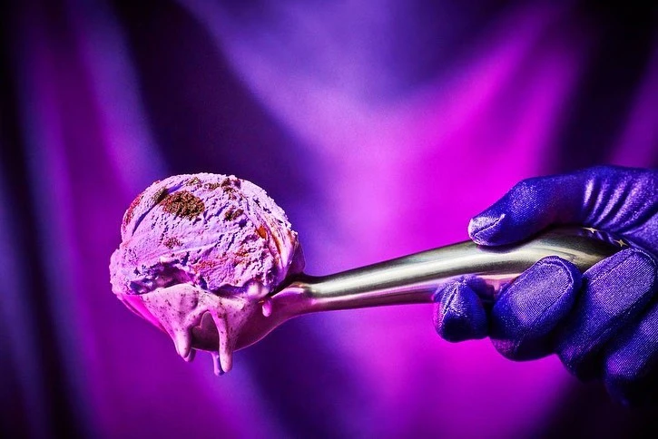 Scoop of lavender ice cream with chocolate flakes on a silver spoon held by a gloved hand, illuminated against a bright purple background.