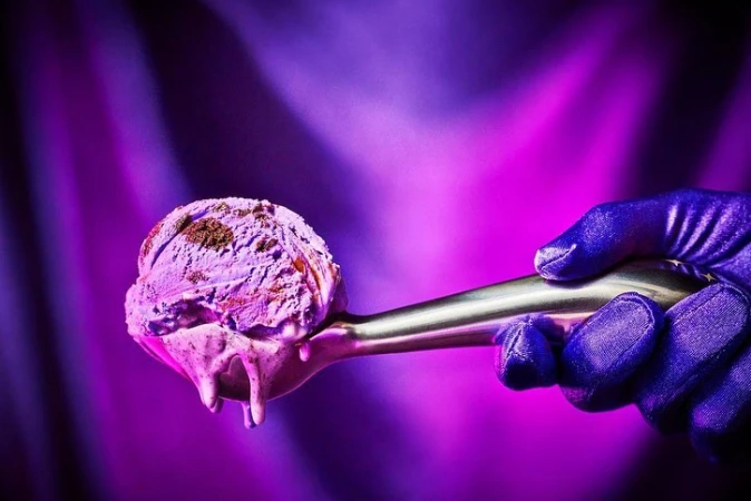 Scoop of lavender ice cream with chocolate flakes on a silver spoon held by a gloved hand, illuminated against a bright purple background.