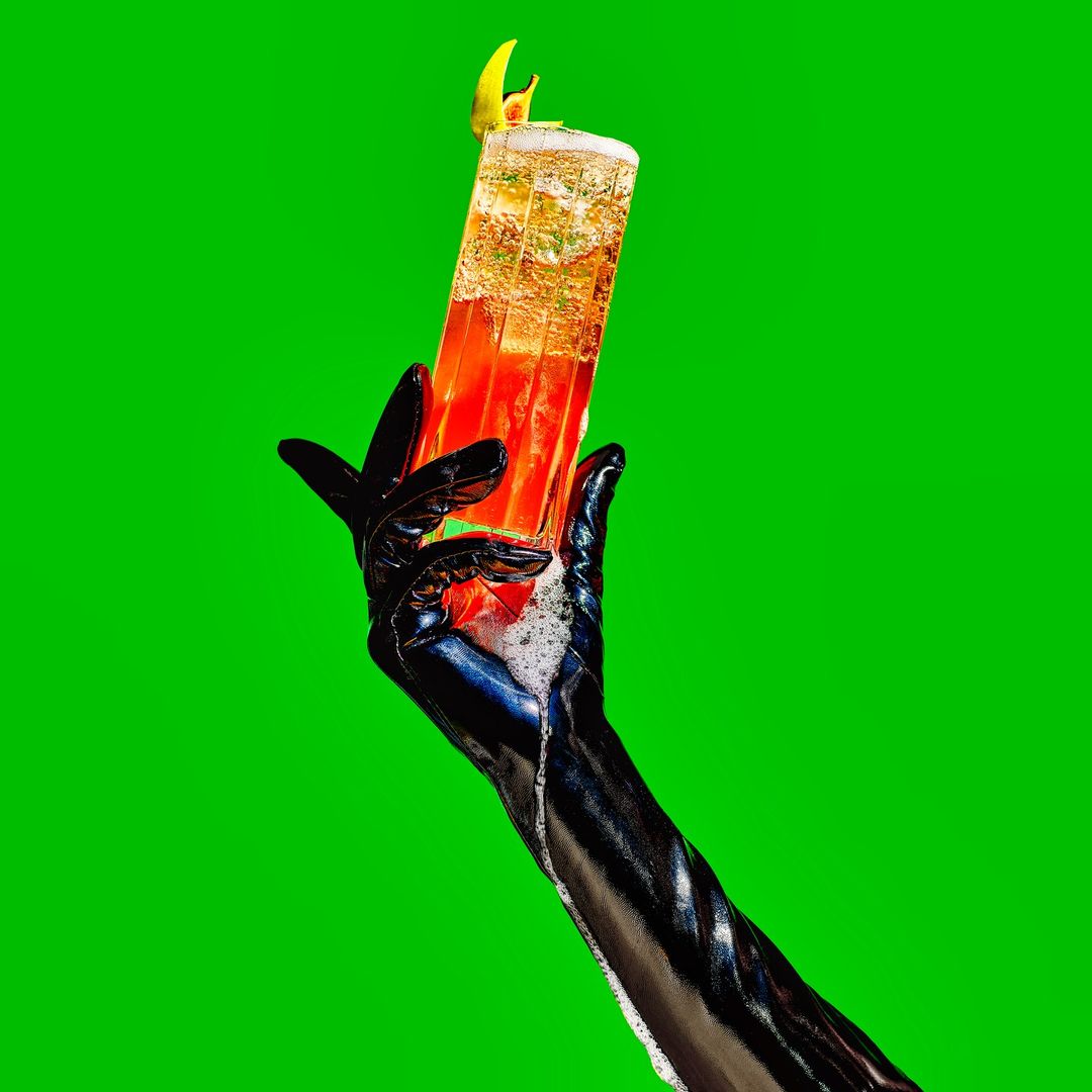 Gloved hand holding a tall glass cocktail with lemon garnish against a bright green background in a studio food and beverage shot.