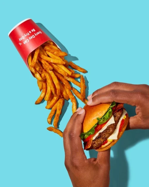 Close-up of a burger and fries held by dark-skinned hands against a turquoise backdrop, suitable for a food and beverage advertisement.