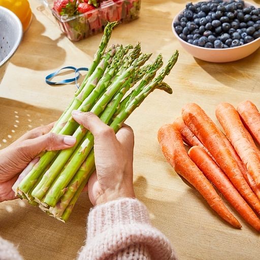 Hands hold fresh asparagus; carrots, blueberries, and strawberries are on a wooden surface, creating a vibrant, healthy food composition.