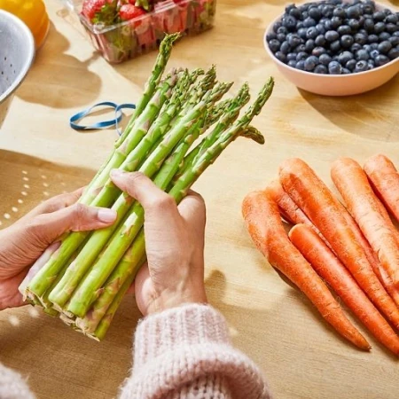Hands hold fresh asparagus; carrots, blueberries, and strawberries are on a wooden surface, creating a vibrant, healthy food composition.