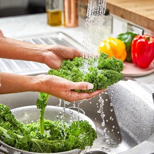 Hands wash fresh green kale under running water in a stainless steel kitchen sink with colorful bell peppers in the background.