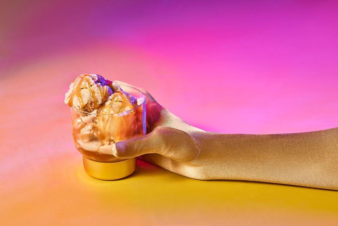 Gloved hand holding a glass of ice cream drizzled with caramel against a vibrant pink and yellow gradient background.