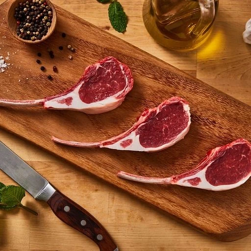 Overhead shot of three raw lamb chops arranged on a wooden cutting board with a knife, spices, oil, and fresh mint sprigs.