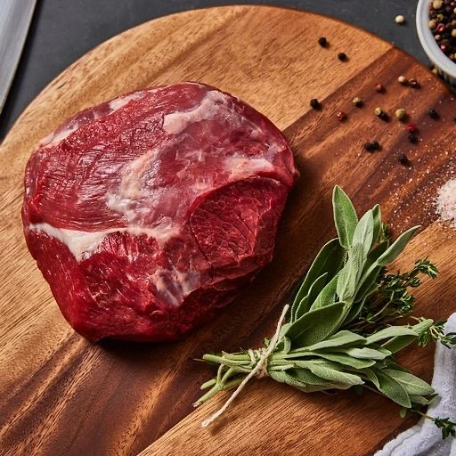 Raw beef roast rests on wooden cutting board alongside fresh herbs, peppercorns, and salt, ready for culinary preparation.