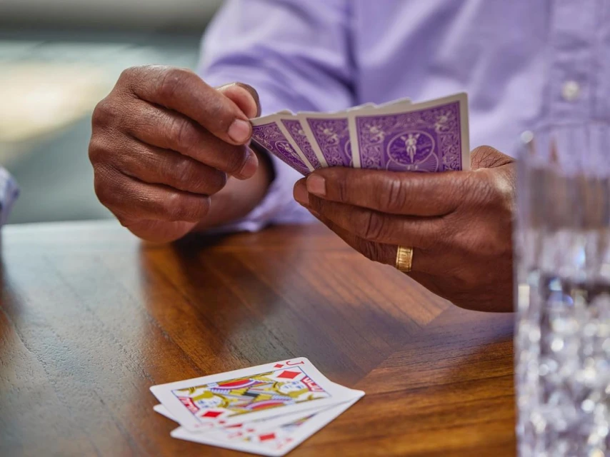 Close-up of a person holding playing cards over a wooden table with a clear glass of water beside the cards.