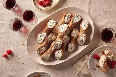 An overhead food photograph of cannoli on a large oval platter with small glasses of red liquid and fresh raspberries on a textured linen cloth.