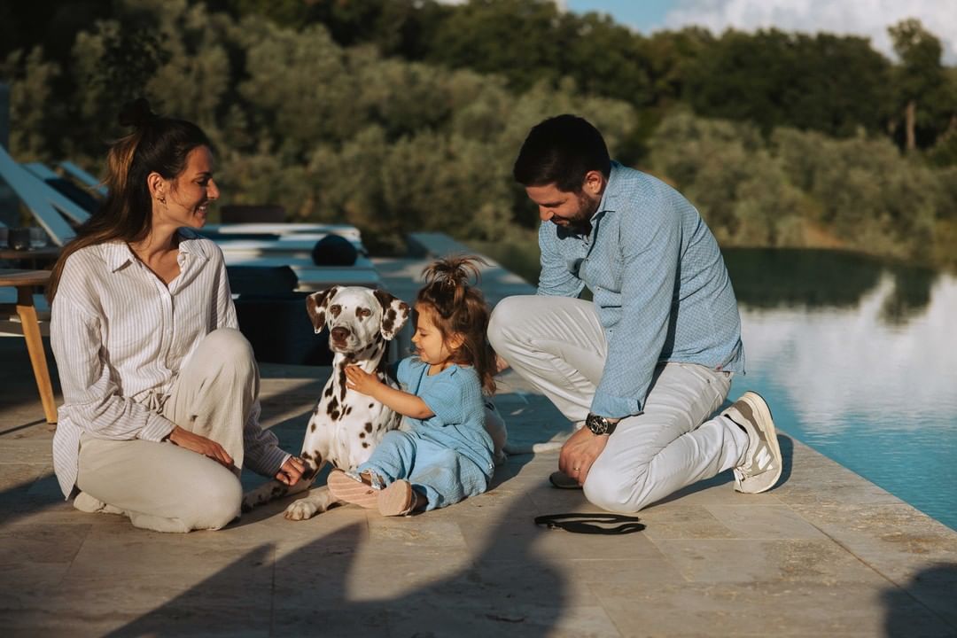 Family with a Dalmatian dog kneeling on a stone patio next to a swimming pool, with greenery in the background on a sunny day.