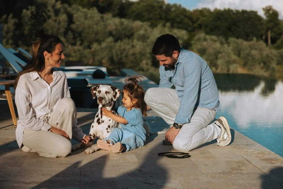 Family with a Dalmatian dog kneeling on a stone patio next to a swimming pool, with greenery in the background on a sunny day.