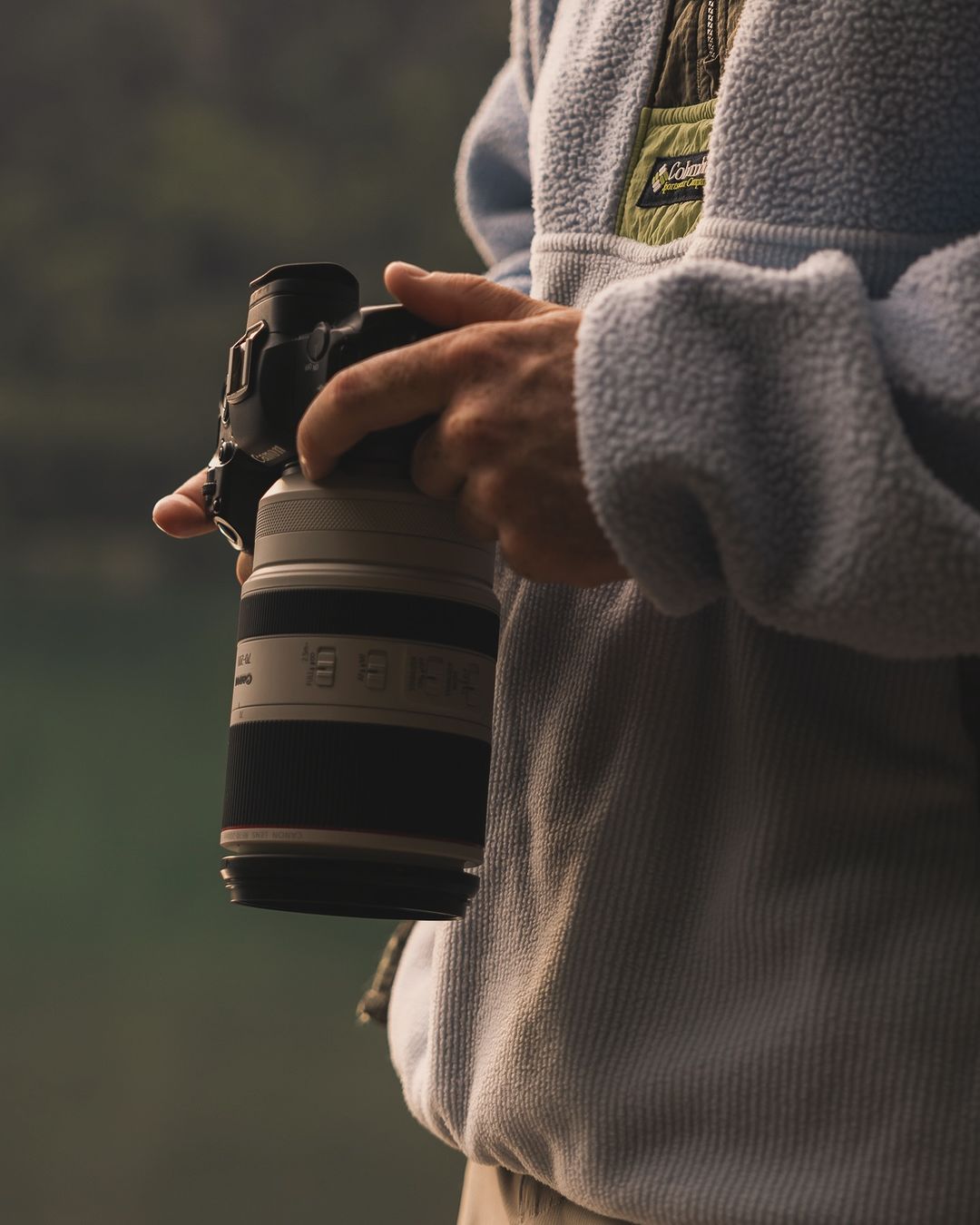 Photographer in fleece jacket holds professional camera with telephoto lens against soft green nature background in outdoor setting.