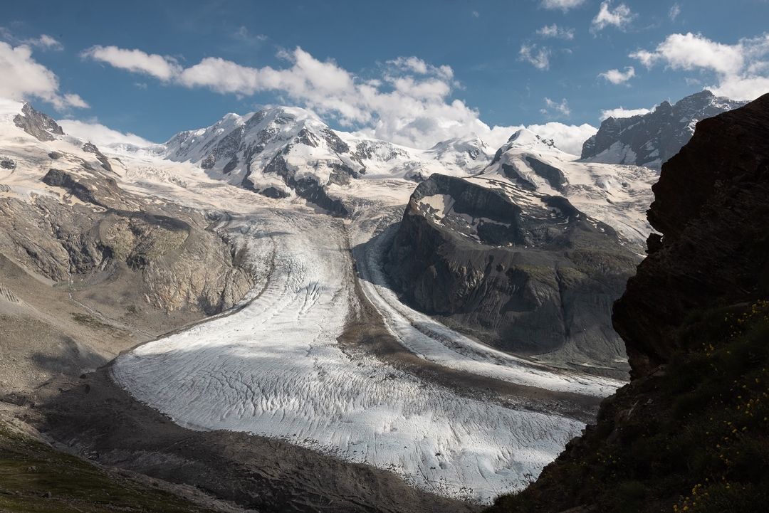 Wide shot of snow-covered mountain range with visible glacier under a partly cloudy sky, framed by rocky outcrop. Alpine landscape.