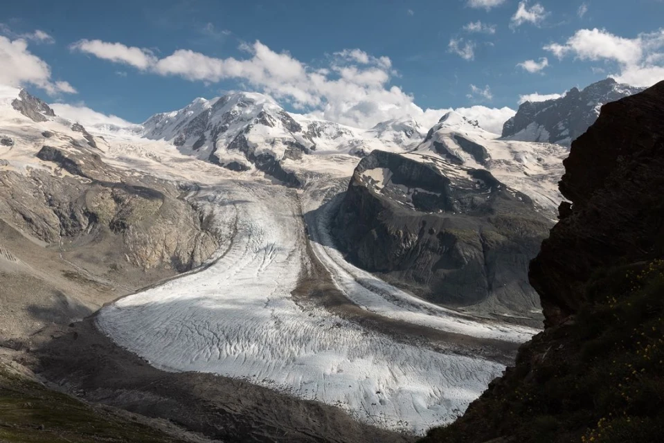 Wide shot of snow-covered mountain range with visible glacier under a partly cloudy sky, framed by rocky outcrop. Alpine landscape.