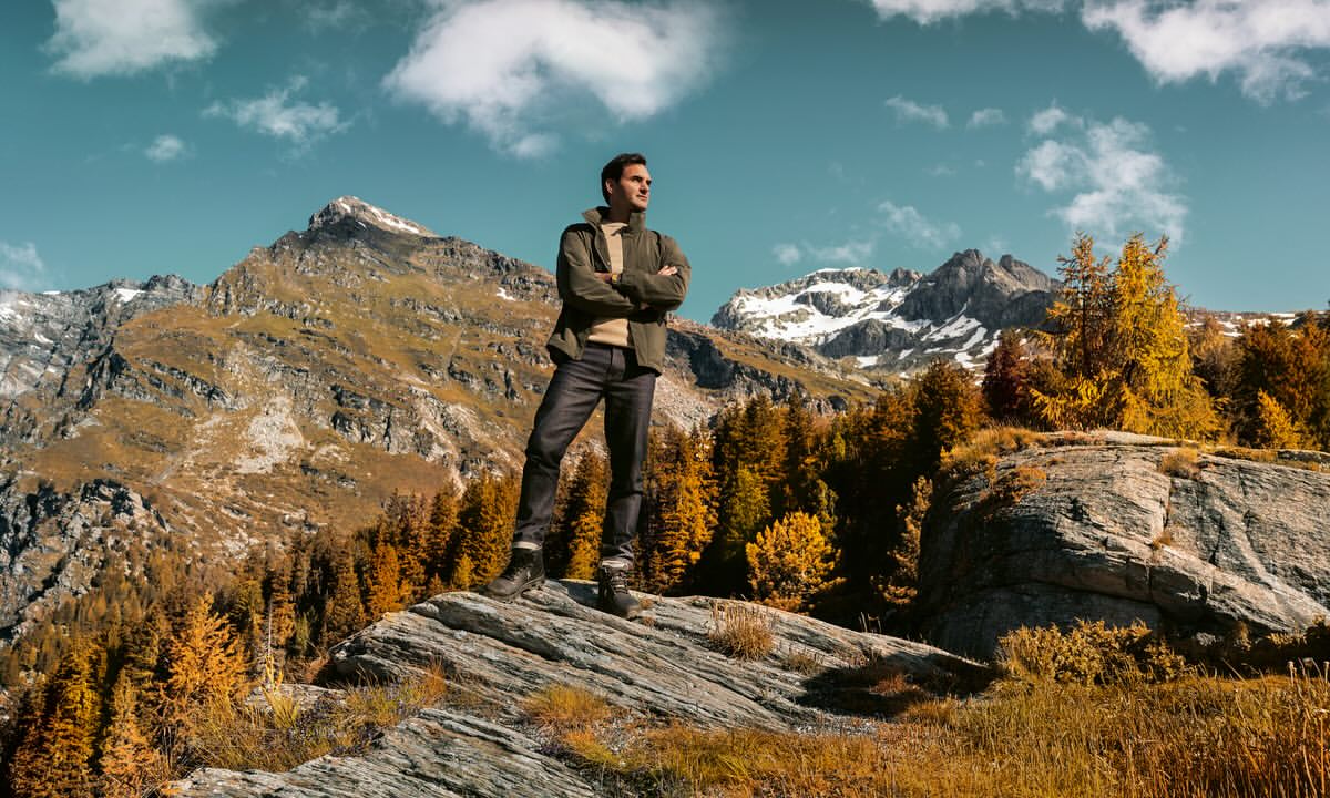 Adult man standing on a rock formation in the mountains, surrounded by autumn trees against a partly cloudy blue sky backdrop.
