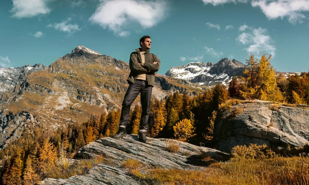 Adult man standing on a rock formation in the mountains, surrounded by autumn trees against a partly cloudy blue sky backdrop.