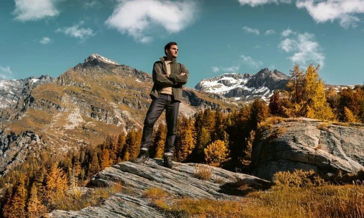Adult man standing on a rock formation in the mountains, surrounded by autumn trees against a partly cloudy blue sky backdrop.