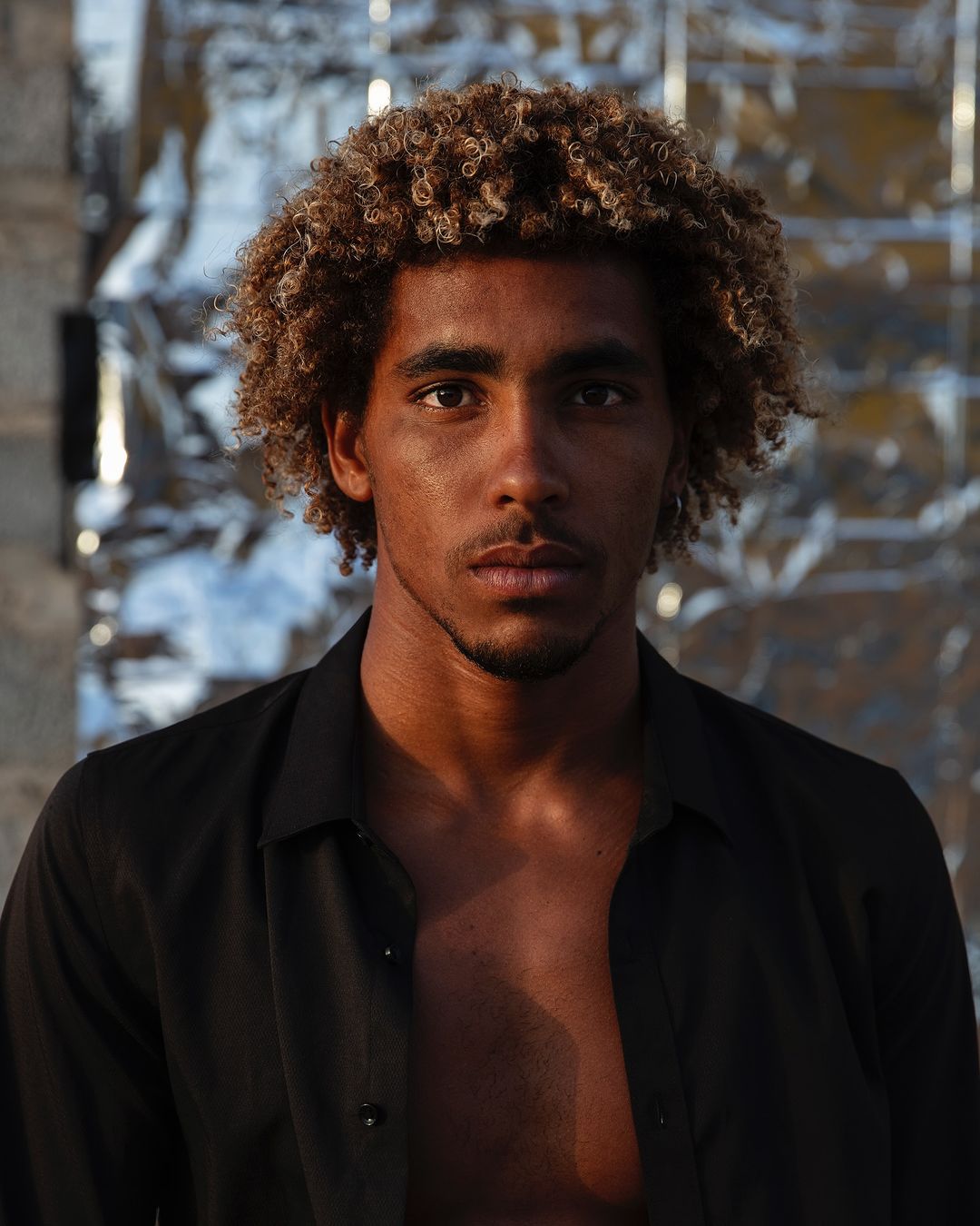 Close-up studio portrait of a young adult male with curly hair. He wears a black, partially unbuttoned shirt against a crinkled foil backdrop.