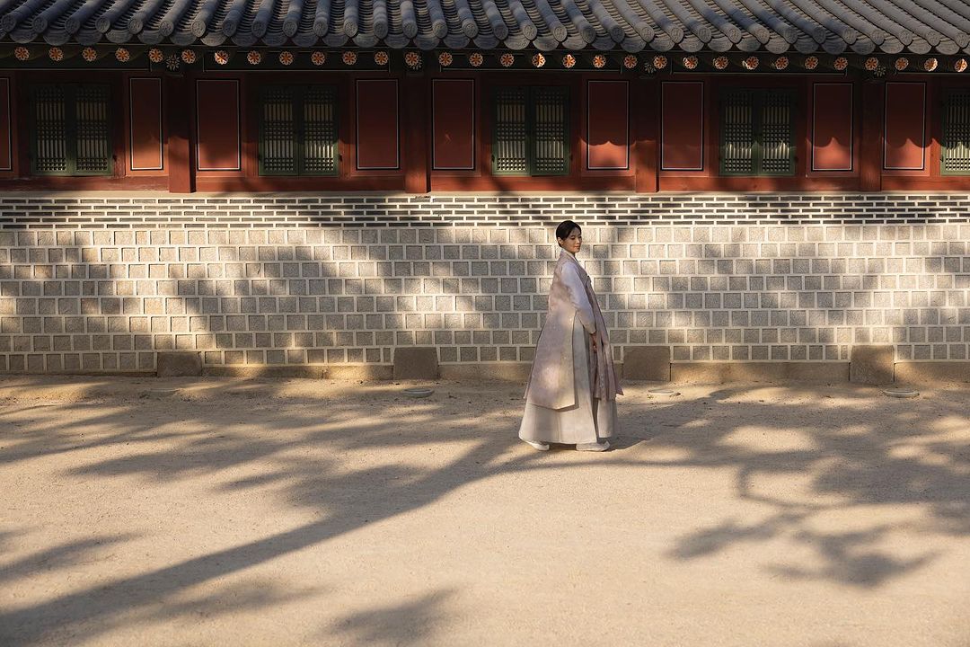 Young woman in a Korean hanbok poses gracefully in front of a traditional building with shadow patterns cast on the stone wall.