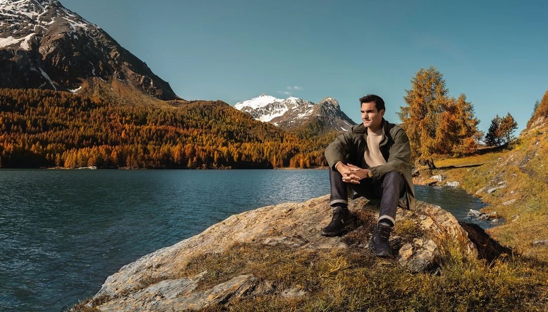 Roger Federer sits on a rock with a lake and mountains in the background. He wears a jacket, jeans, and hiking boots in nature.