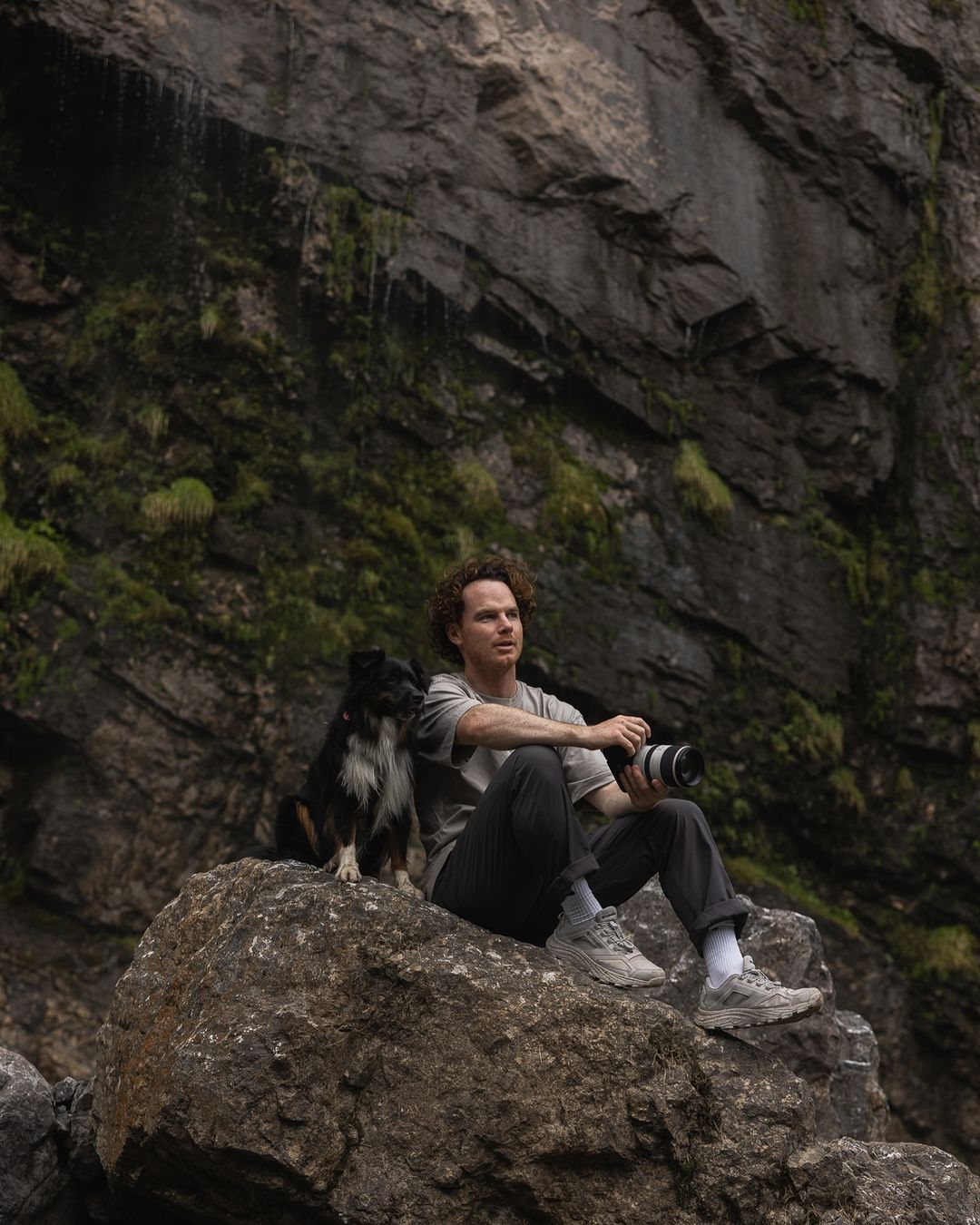 Man and dog sit on a rock in front of a rock cliff. The man holds a camera. The setting is outdoors with rocky and green foliage.